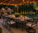 Modern outdoor dining set beneath a wooden pergola with string lights, a stone fireplace, and beautifully landscaped backyard.